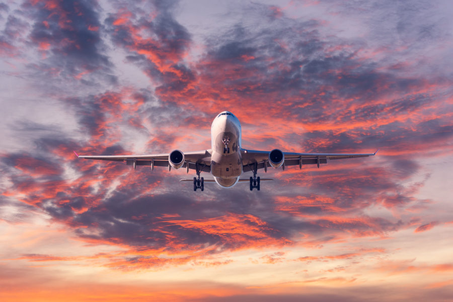 Landscape with passenger airplane is flying in the blue sky with red, purple and orange clouds at sunset.